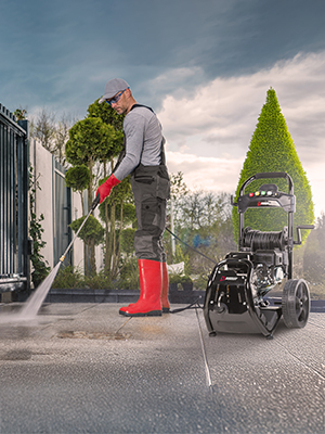 Person in dark coat and red boots trimming a conical topiary bush with electric trimmer. Wheelchair visible nearby, cloudy sky in background.