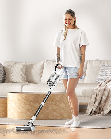 Person in white t-shirt and shorts using cordless vacuum cleaner in living room with beige couch and pillows.