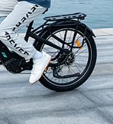 Vista parcial de la rueda trasera, el guardabarros y el cuadro de una bicicleta eléctrica contra una superficie de concreto. Neumático negro con radios plateados visibles.