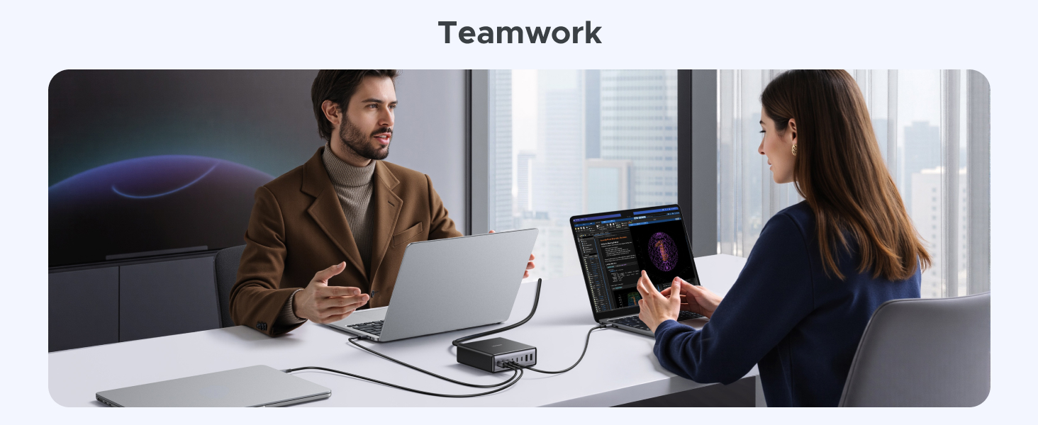 Two people collaborating at a desk with laptops and tablets. One person gestures while explaining, showcasing teamwork in a modern office setting with city views.