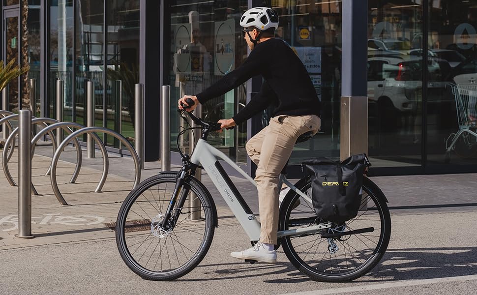 Urbane Straßenszene mit einer Person, die mit einem schwarzen Elektrofahrrad an Ladenfronten und Fahrradständern auf dem Bürgersteig vorbeifährt.