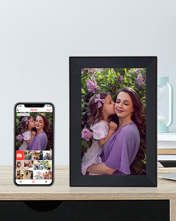 Digital photo frame displayed on wooden surface next to smartphone showing photo gallery interface.