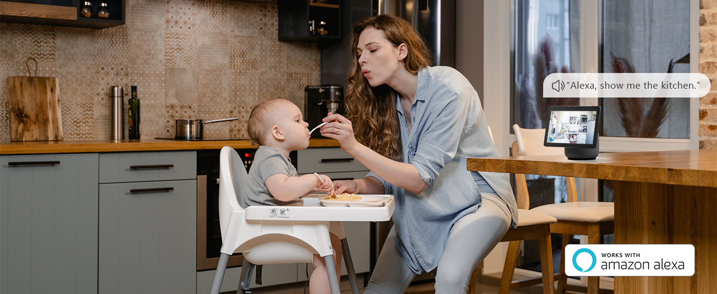 Kitchen scene with a person feeding a baby in a high chair. A smart display is visible on the counter in the background.