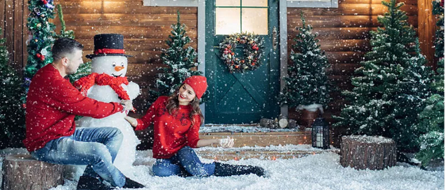 Winterszene mit Familie und Schneemann. Weihnachtsbäume, Kranz und fallender Schnee sichtbar. Menschen in roter Kleidung sitzen auf verschneitem Boden.