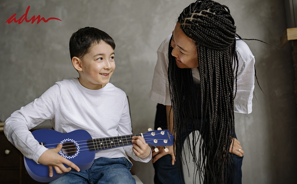 A young boy plays a blue acoustic guitar while an older person with braids leans over, interacting with him. Both are smiling in a casual indoor setting.