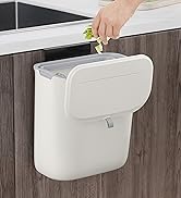 Wall-mounted kitchen compost bin with lid. White plastic container attached to cabinet, hand dropping vegetable scraps inside.