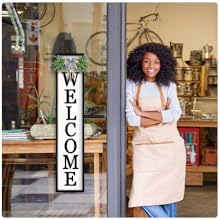 A woman in apron smiles by a glass door with a &#34;WELCOME&#34; sign and maple leaf.