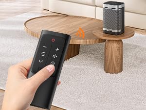 Portable space heater on wooden coffee table with hand holding remote control. Heater has metallic grille design. Remote features multiple buttons for temperature and settings control.
