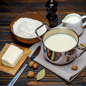 Ingredientes para cocinar sobre una superficie de madera: tazón de harina, olla de leche, mantequilla, molinillo de pimienta, jarra de crema, cuchillo y frutos secos para untar. Demuestra preparación para hornear o cocinar.