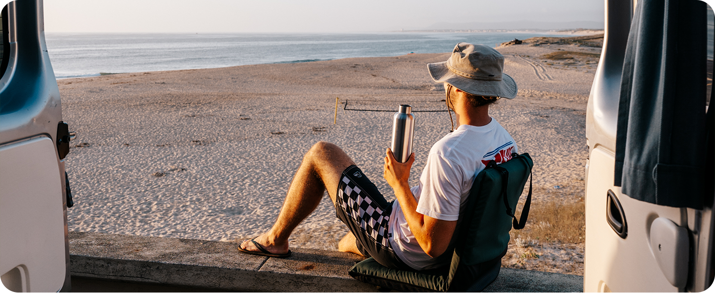 Person sitting in vehicle looking out at desert or beach landscape through open door, wearing casual attire and hat.