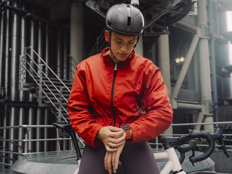 Persona con chaqueta roja resistente a la intemperie y casco de seguridad que comprueba en la muñeca en un entorno industrial urbano con estructuras metálicas en el fondo.