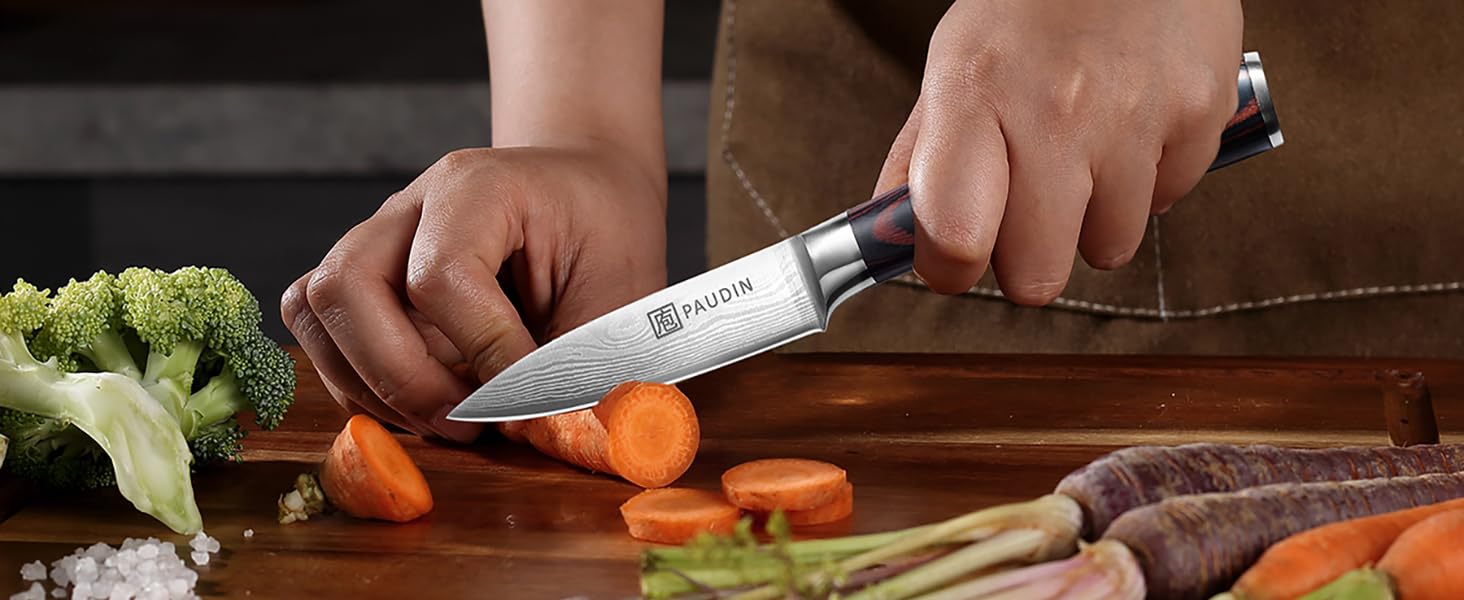 Chef's knife in use, cutting carrots on wooden board. Hands visible holding knife and carrot, with various vegetables nearby.