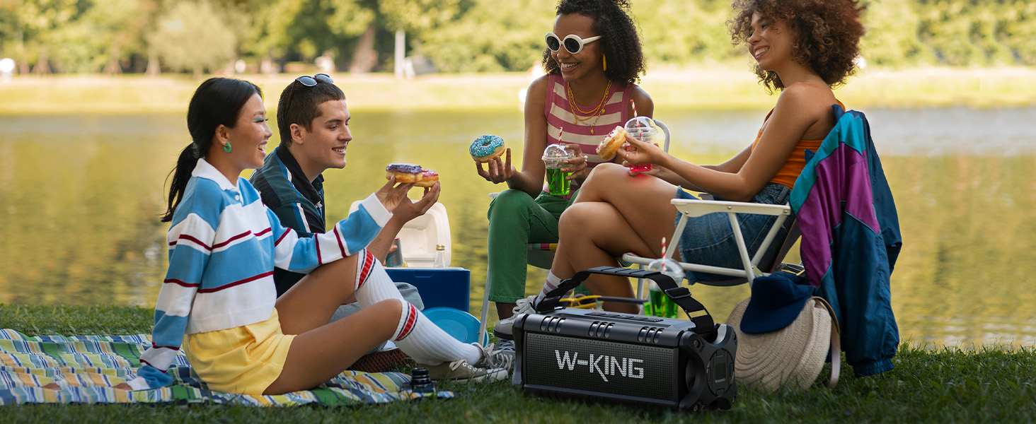Group enjoying outdoor picnic with beverages by a lake, sitting on blankets with a cooler nearby.