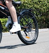 Primer plano del pie de un ciclista sobre el pedal de una bicicleta, con un calcetín deportivo blanco y mostrando una rueda parcial en