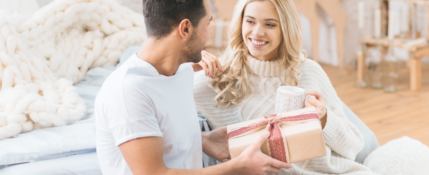 A couple sitting on a couch, exchanging a gift. The woman is smiling while receiving a wrapped present from the man.