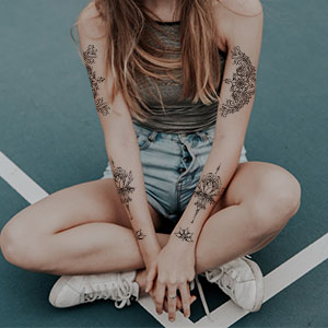Persoon die met gekruiste benen op een gevoerd oppervlak zit, witte sneakers, een spijkerbroek en een donkere top draagt. Meerdere bloementattoos zichtbaar op de armen.