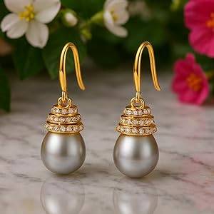 Gold dangle earrings featuring large gray pearls with ornate tiered gold caps set with small gemstones. Earrings displayed on marble surface with colorful flowers in background.