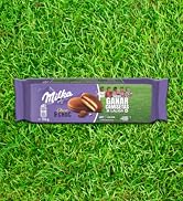 El texto dice «Milka». El envase de la barra de chocolate morado se muestra sobre un fondo de hierba, con imágenes de productos de chocolate y galletas.