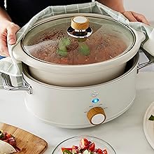 Overhead view of round electric slow cooker or warming dish with food inside. Additional plates with garnishes visible on white surface.