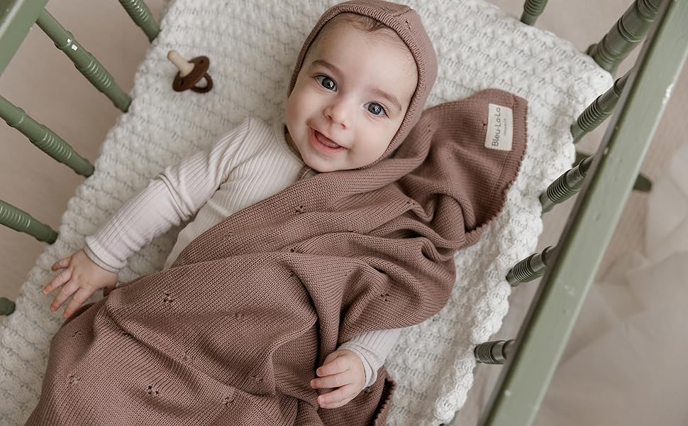 Knitted baby clothing in soft brown tones displayed on white textured bedding.