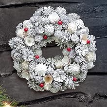 a white christmas wreath with pinecones and red berries on a black wooden wall.