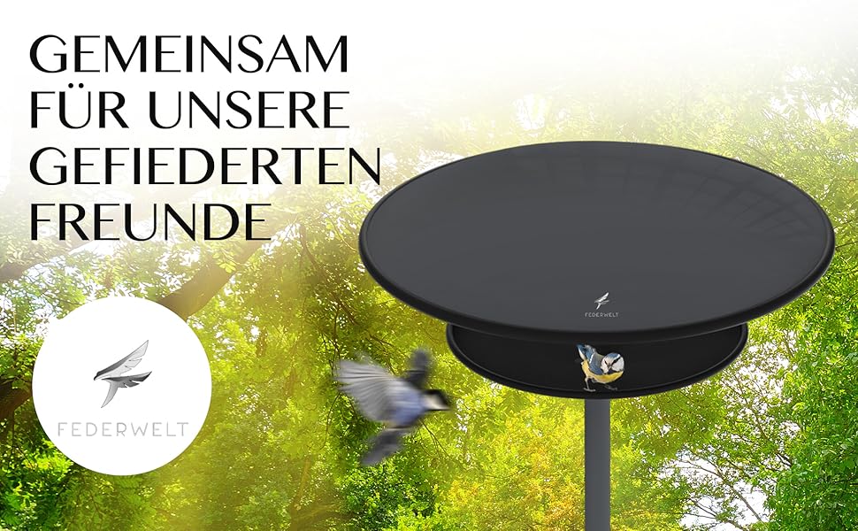 Schwarze, runde Vogelfutterplattform mit erhöhtem Rand, montiert auf einer Stange. Der Hintergrund zeigt verschwommenes Grün und Text auf Deutsch.