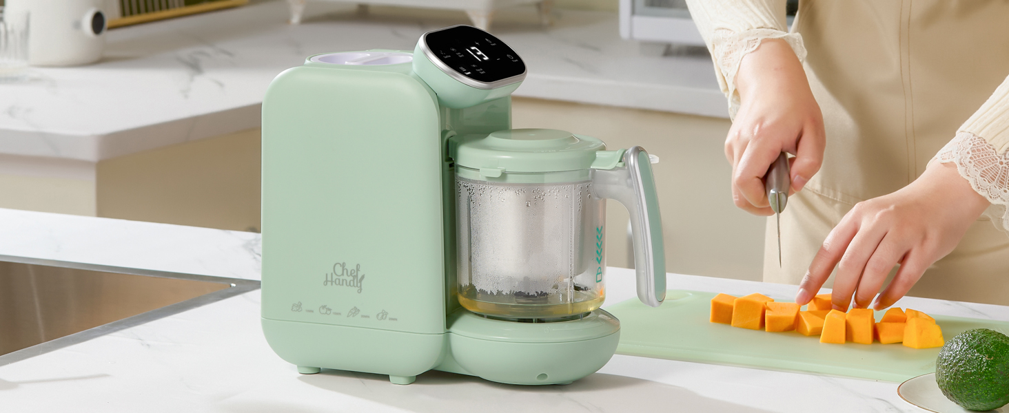 Mint green baby food maker with digital display, blending container, and steaming basket. Shown in kitchen setting with person cutting vegetables nearby.