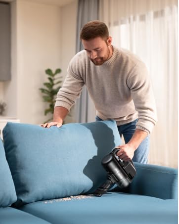 Person, die eine blaue Couch mit einem Handstaubsauger in einem gut beleuchteten Raum mit Zimmerpflanze im Hintergrund reinigt oder pflegt