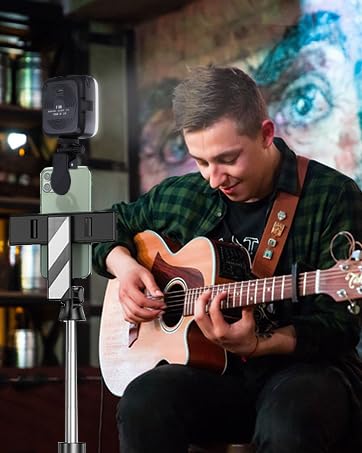 Person playing acoustic guitar while seated, with small camera mounted on tripod in foreground. Colorful mural visible in background.