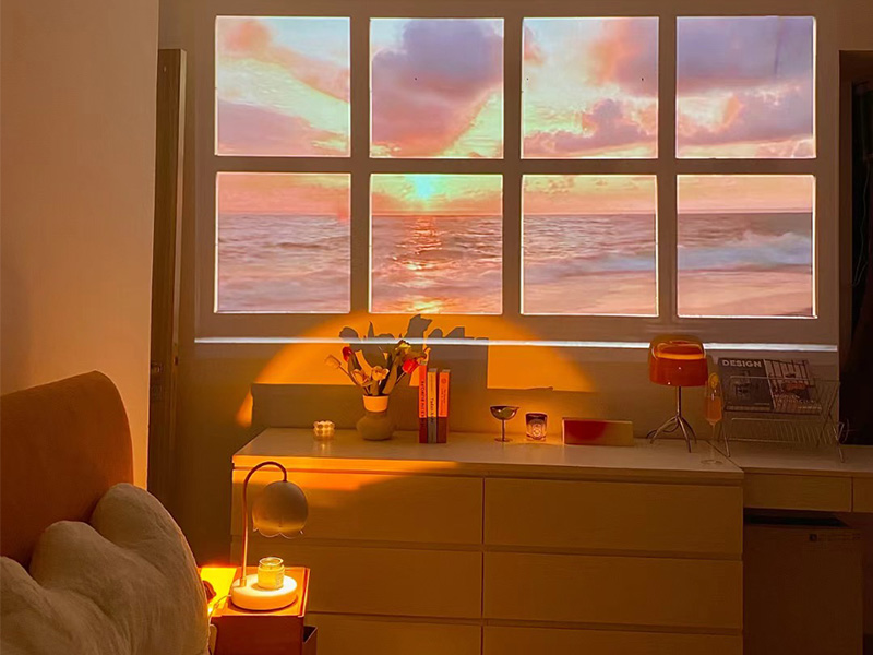 Intérieur de chambre confortable avec vue sur le coucher de soleil sur l'océan depuis la fenêtre. Commode avec objets décoratifs et lampe, fauteuil partiellement visible au premier plan.