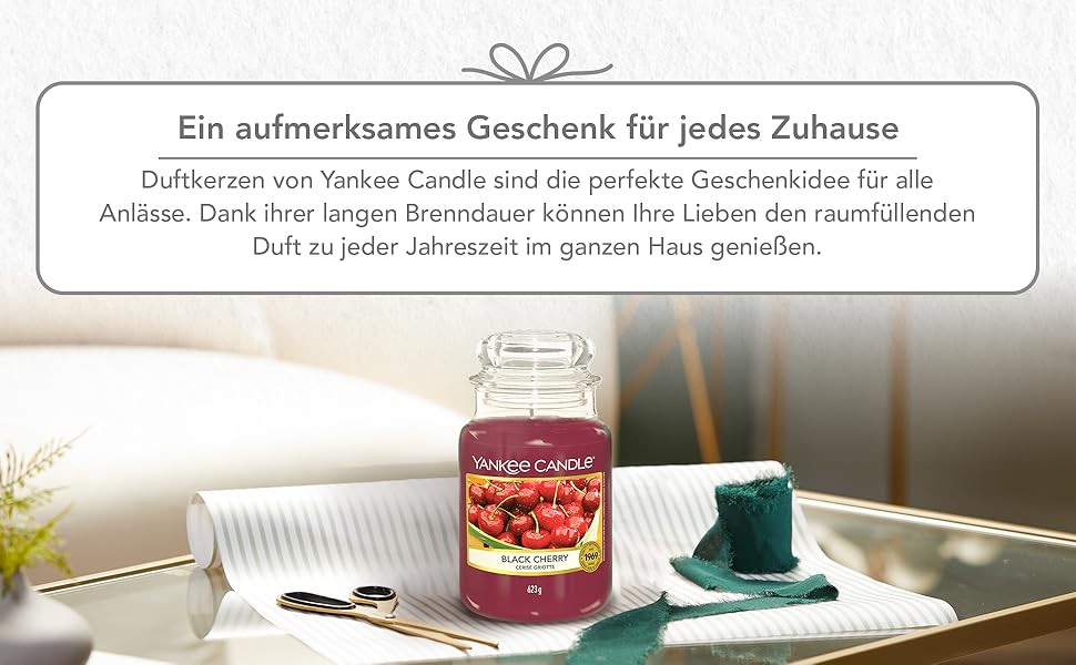 Text: „Ein entspannendes Geschenk für jedes Zuhause“ mit roter Glaskerze und blaufarbener Tasse auf heller Oberfläche