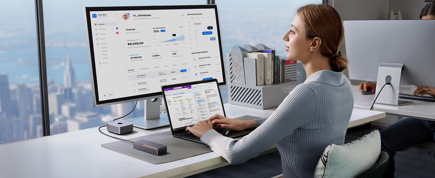 Femme au bureau utilisant une configuration informatique à double écran avec grand écran et ordinateur portable. Les toits de la ville sont visibles à travers la fenêtre.