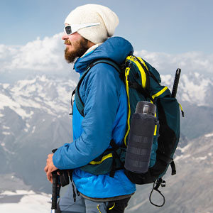 Excursionist with blue jacket and hat, black backpack with yellow straps, standing on the snowy top of a mountain with views of