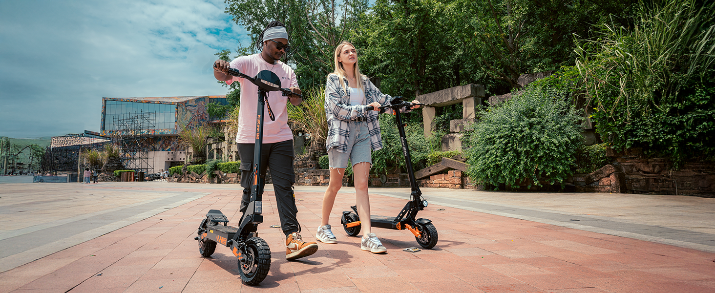 Due persone in sella a scooter elettrici su un sentiero asfaltato all'aperto. Gli scooter hanno ruote e manubri di grandi dimensioni. Sullo sfondo sono visibili alberi ed edifici