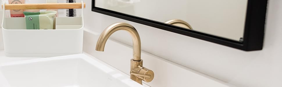 Gold-finished bathroom faucet with curved spout and white organizing tray containing personal care items visible in background.