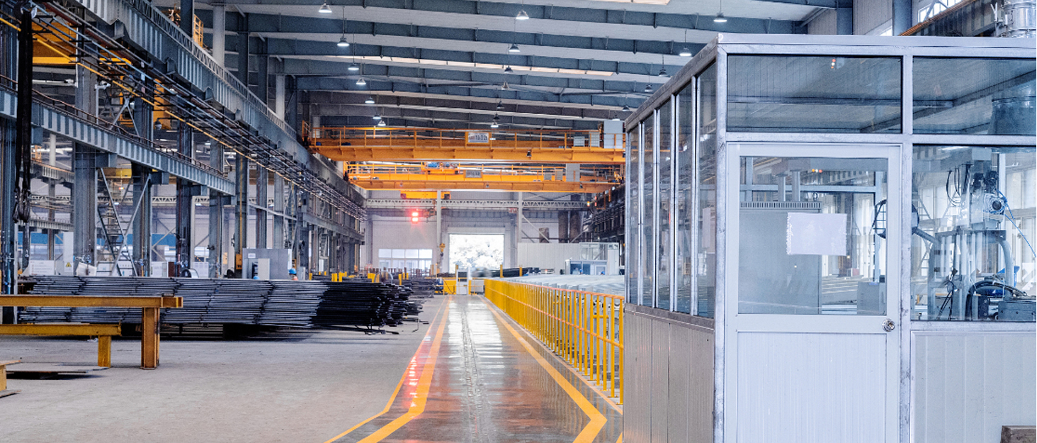 Industrial warehouse interior with overhead crane, metal sheets stacked on floor, yellow safety barriers, and a glass-walled office area. High ceilings with bright lighting illuminate the spacious facility.