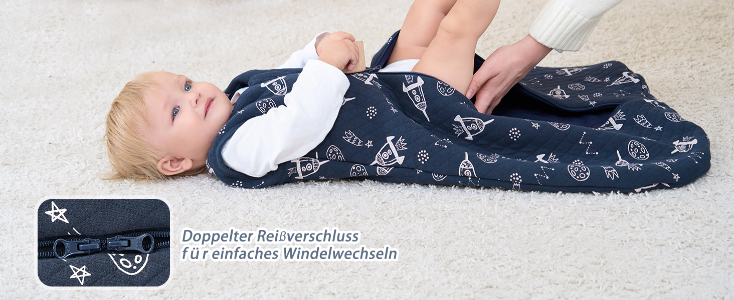Säugling liegt auf einem marineblauen Schlafsack mit weißem Muster. Der Schlafsack verfügt über einen Reißverschluss und ist als „Doppelter Reiseschlafsack“ (doppelter Reiseschlafsack) gekennzeichnet