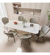 Modern dining set with white marble table and six beige upholstered chairs. Built-in shelving displays small appliances. Bowl of tomatoes and plant centerpiece on table.