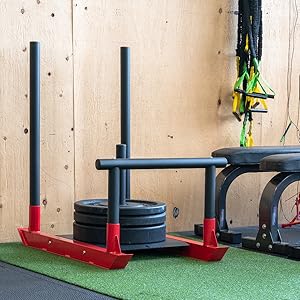 Loaded red power sled at an angle, push poles against plywood wall, on green turf, in gym
