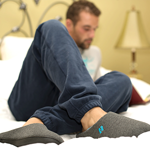 Man sitting on bed in the bedroom, reading a book and relaxing in a pair of RockDove slippers.